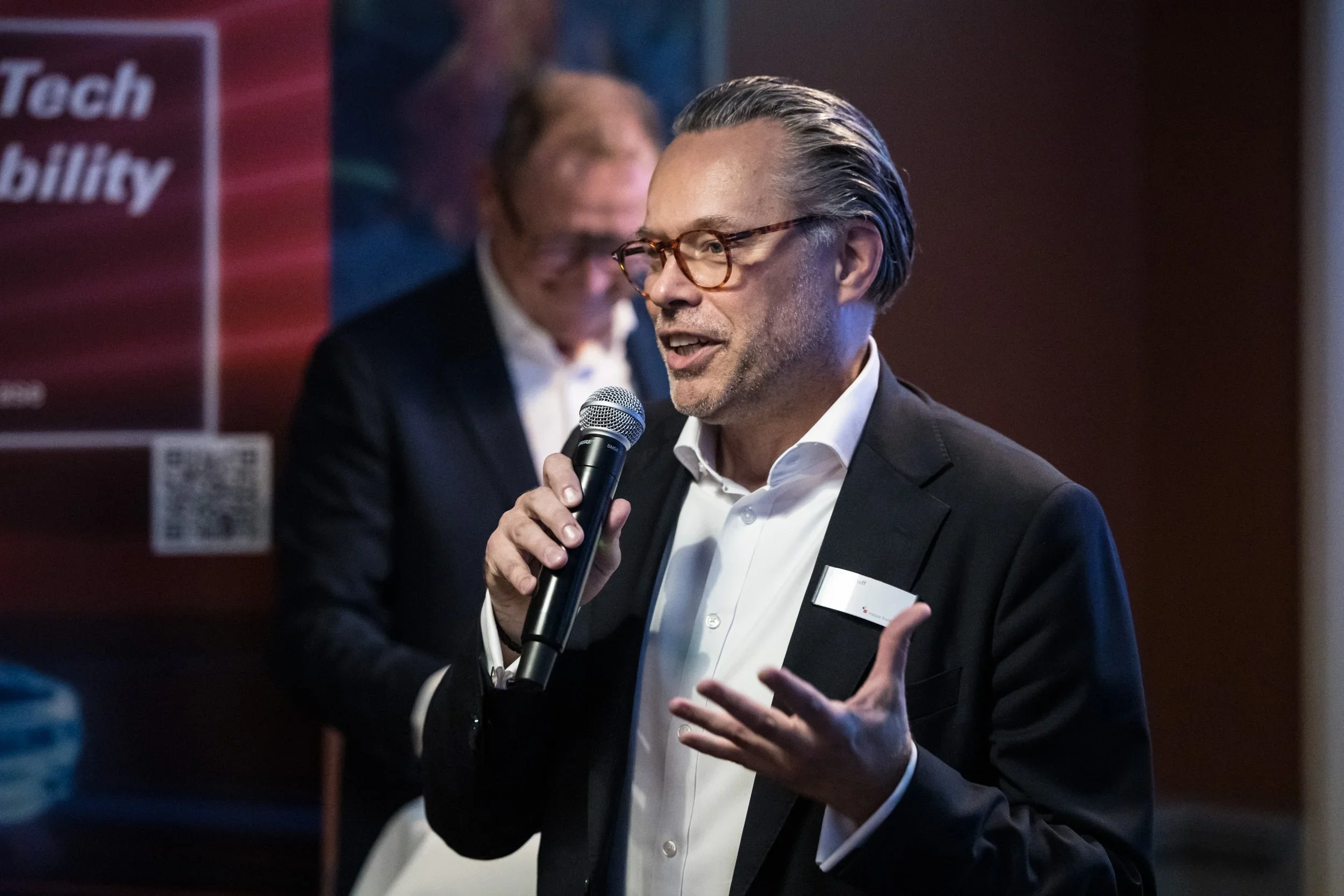 A man in a black suit and glasses speaking into a microphone at a conference or event.