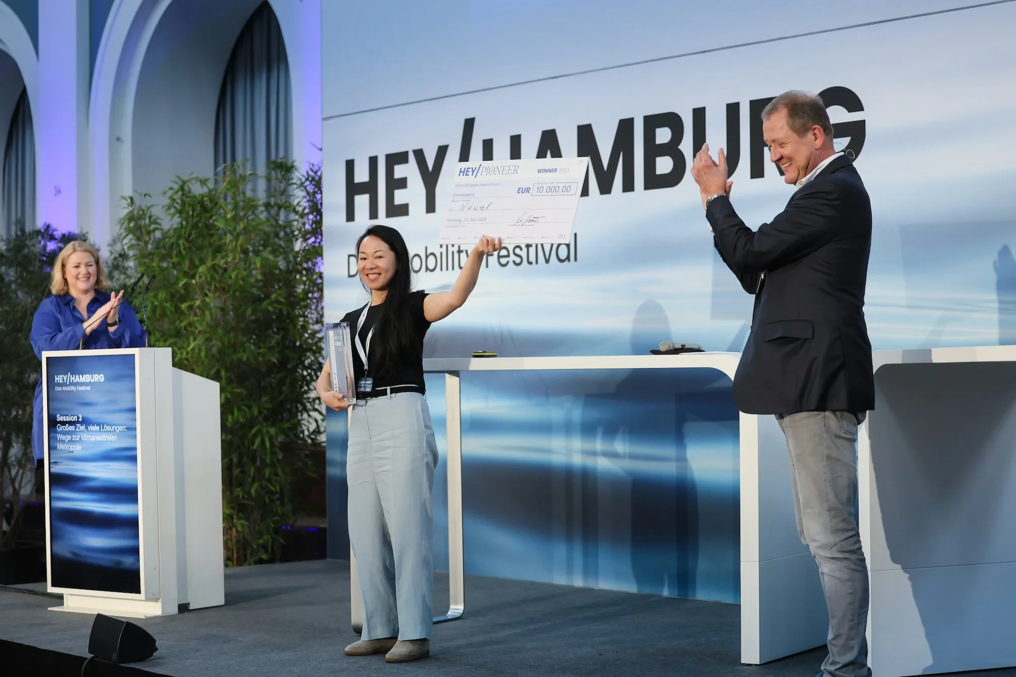 A woman on stage holding large check with €10,000 at the Hey Hamburg mobility festival, smiling as a man claps beside her.