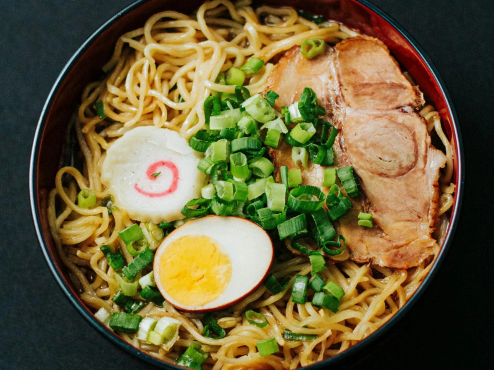 Bowl of ramen with sliced pork, chopped green onions, half a boiled egg, fish cake with a red swirl, and noodles.