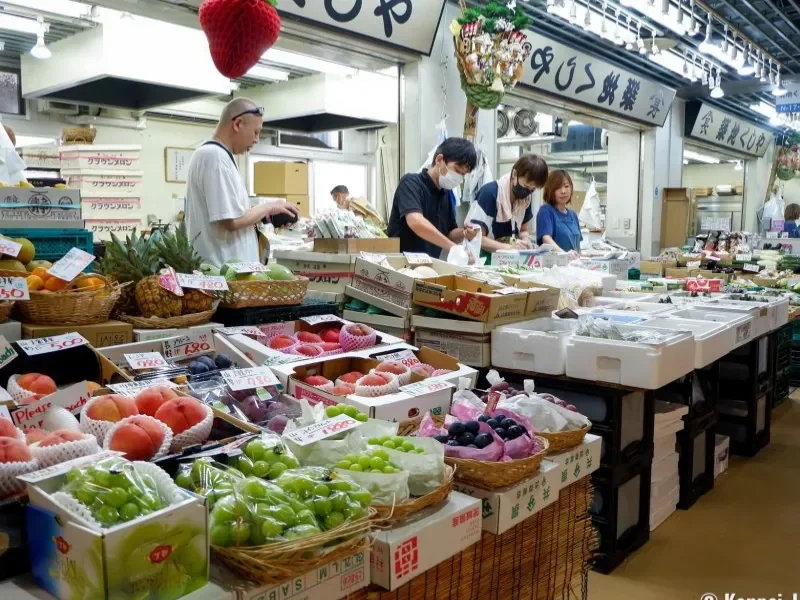 Fresh fruits including peaches, grapes, and blueberries displayed at a market stall, with vendors and customers shopping.