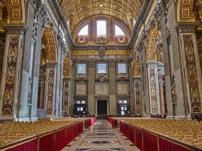 Interior of a grand, ornate church or cathedral with gilded decorations, tall columns, and a high, decorated ceiling. Rows of chairs are lined up along the aisle with red cloths.