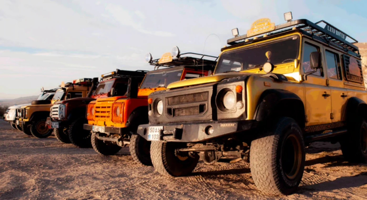 A lineup of off-road vehicles parked on a dirt terrain under a partly cloudy sky.