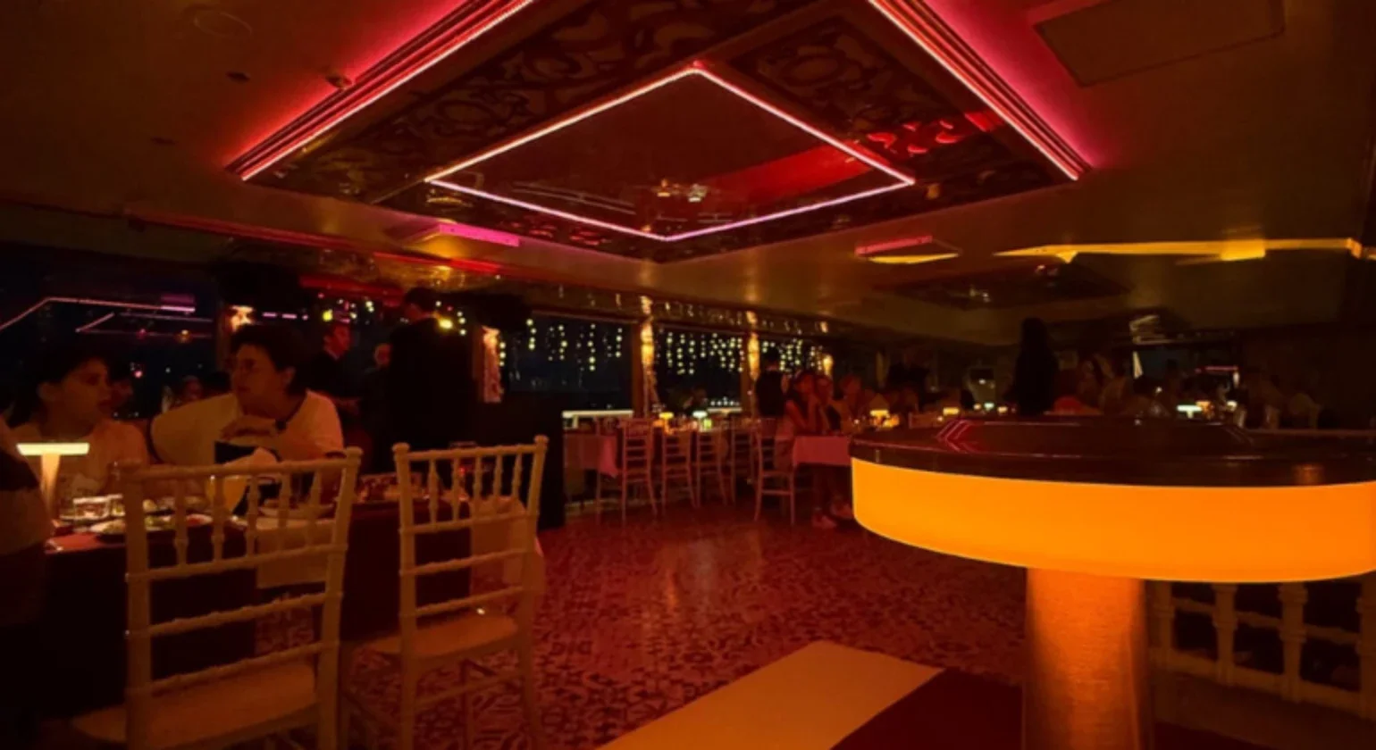 Dimly lit restaurant or lounge with people seated at tables, illuminated by warm, colorful lights, and mirrored ceiling decor.