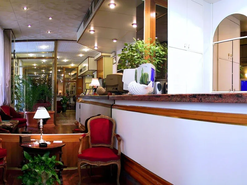 Interior of a cozy lounge area with red upholstered chairs, a small wooden table with a white lamp, potted plants, and a bar counter with decorative items and a computer monitor.