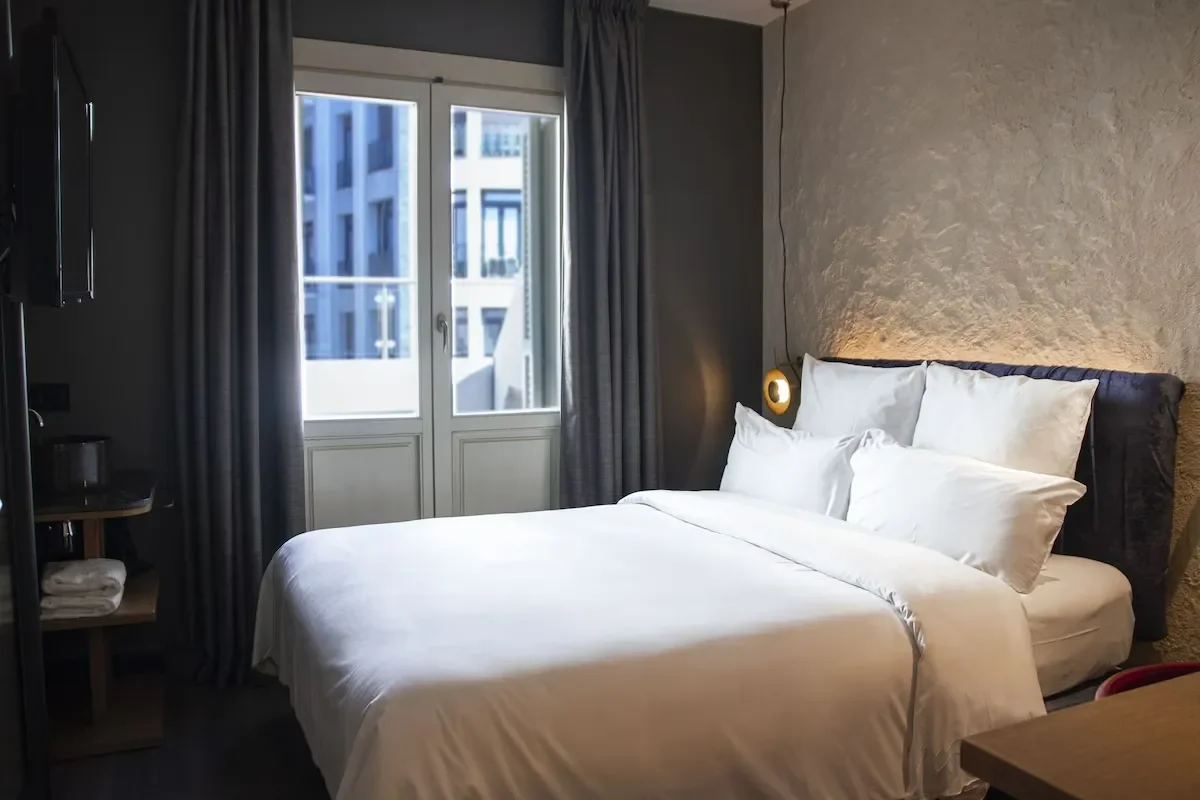 A modern hotel room with a neatly made bed, white bedding and pillows, a bedside lamp, a window with gray curtains, and a view of neighboring buildings.