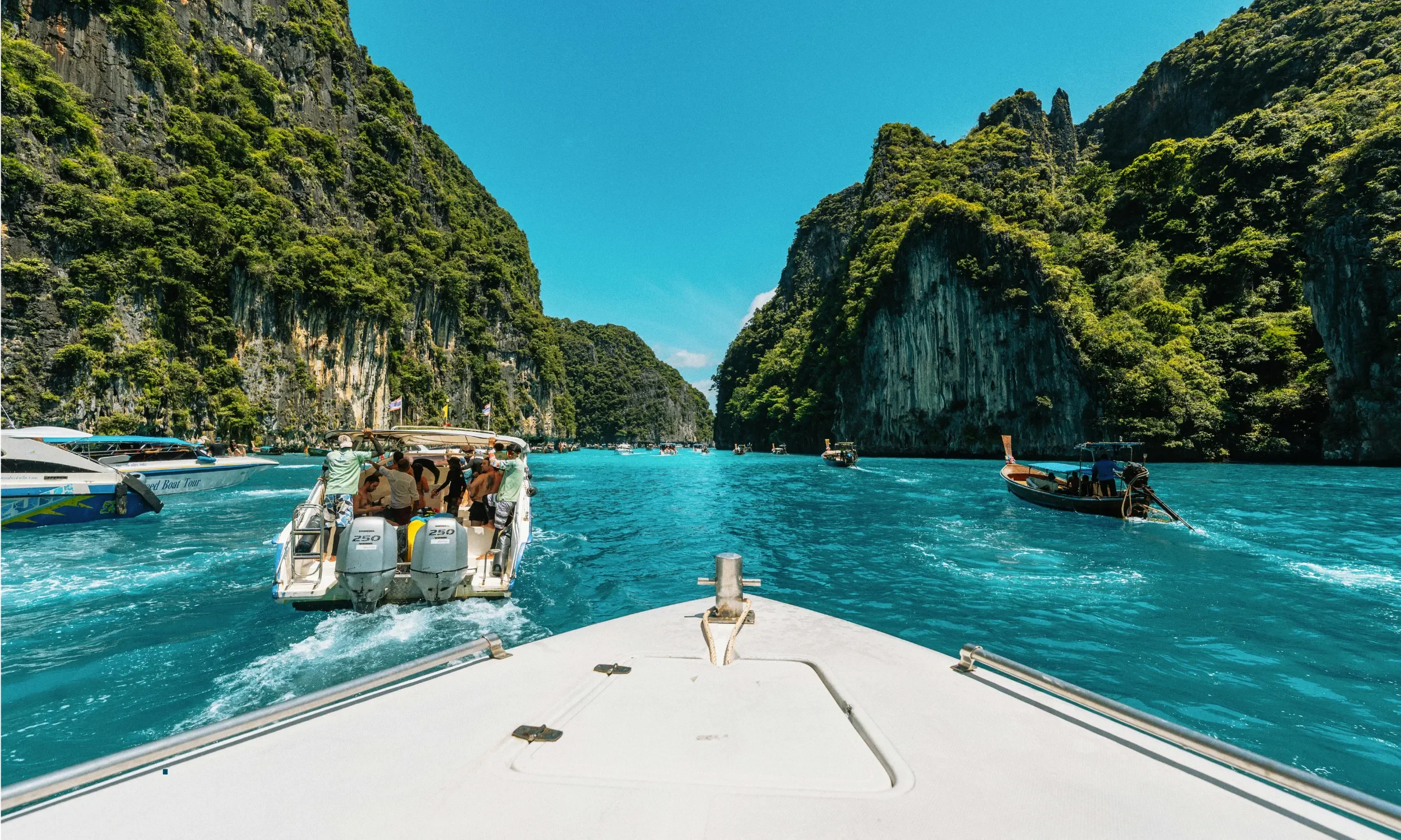 View from the front of a boat looking at turquoise water with multiple boats and limestone cliffs covered in green foliage on either side under a clear blue sky.