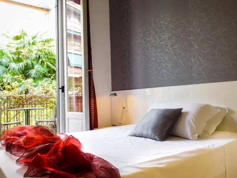 Hotel room with a bed, gray pillow, white sheets, and a red sheer fabric blanket near open balcony door, showing green foliage outside.