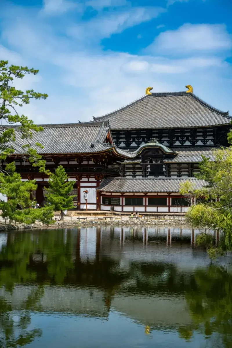 Traditional Japanese temple with black and white walls, curved roofs with golden ornaments, surrounded by green trees, and reflected in a pond under a partly cloudy sky.