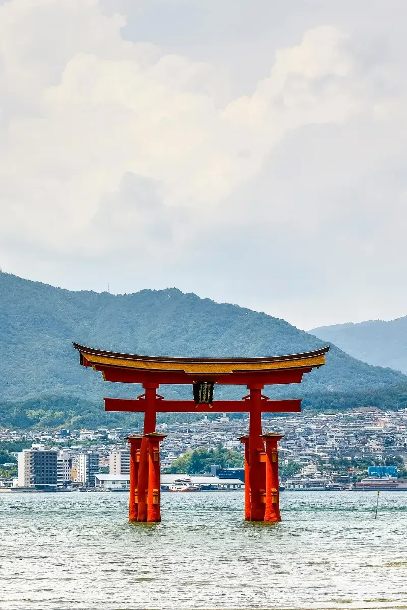 A large red torii gate standing in water with a city and mountains in the background.