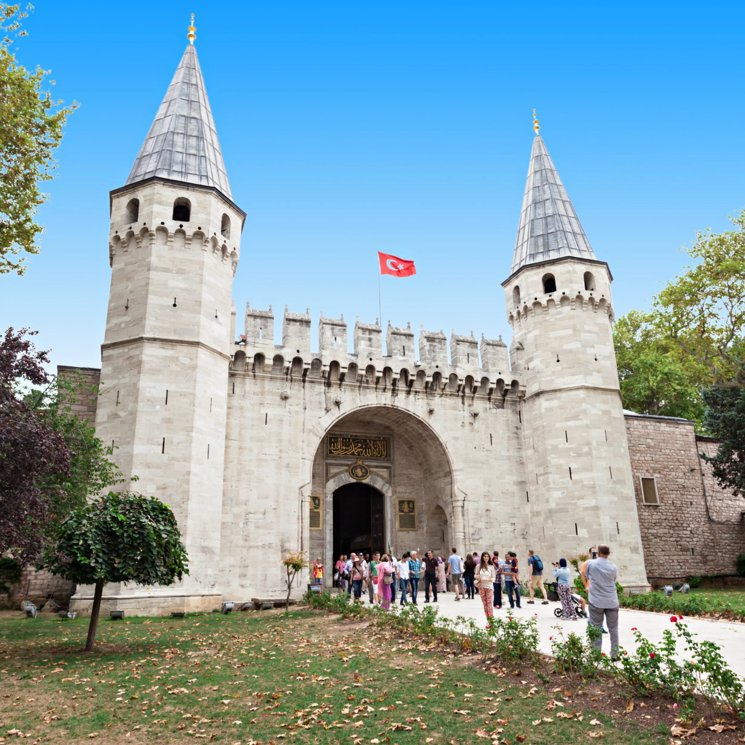 Historic stone castle gates with two tall pointed towers and a Turkish flag flying between them, surrounded by a courtyard with people walking and trees with autumn leaves.