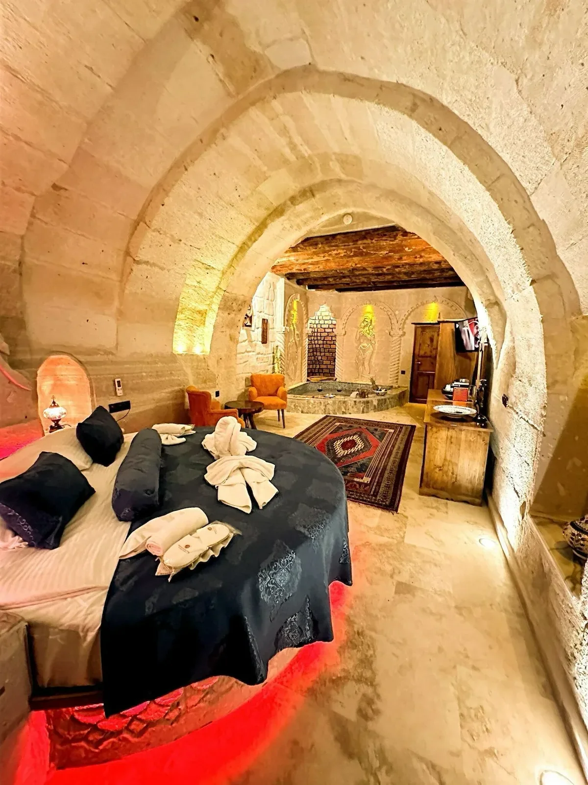 A cozy basement room with stone arches, a large round bed with black and pink bedding, folded towels, a small seating area with two orange chairs, a rug, a stone hot tub, and wooden furniture, illuminated by warm lighting.