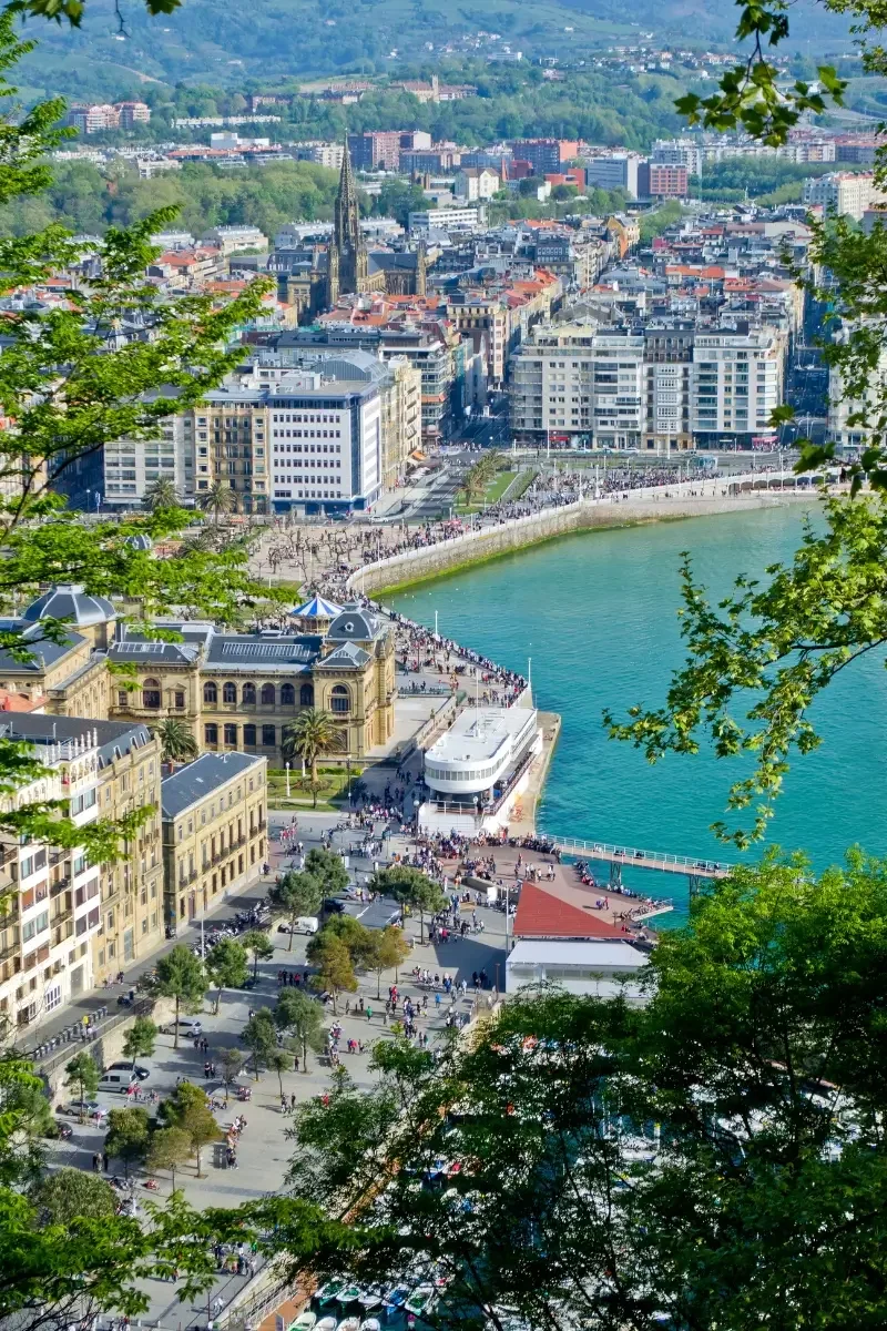 A panoramic aerial view of a city with a river, historic buildings, and a crowd walking along the waterfront. The cityscape includes a prominent church with a tall steeple and surrounding green hills.