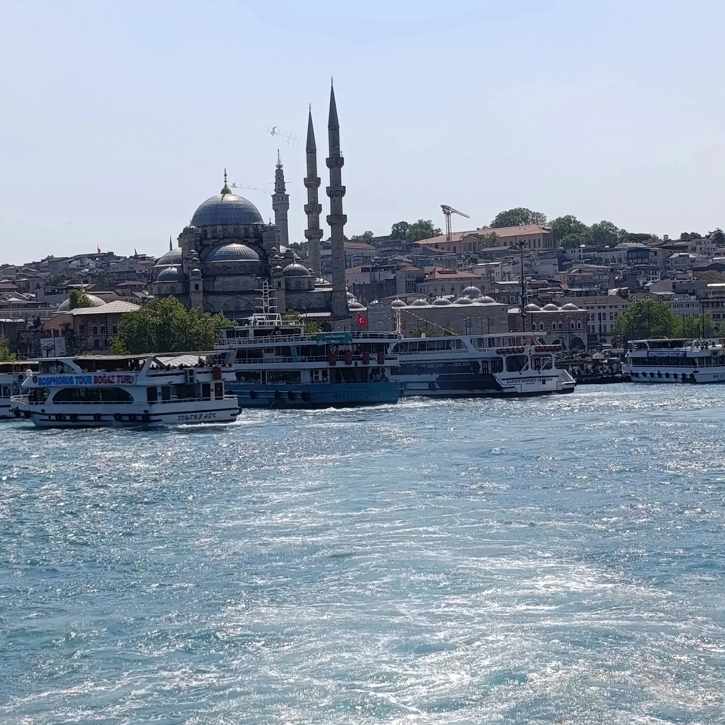 A view of the Bosphorus Strait with boats sailing, and a cityscape with a mosque featuring multiple domes and minarets in the background, under a clear blue sky.