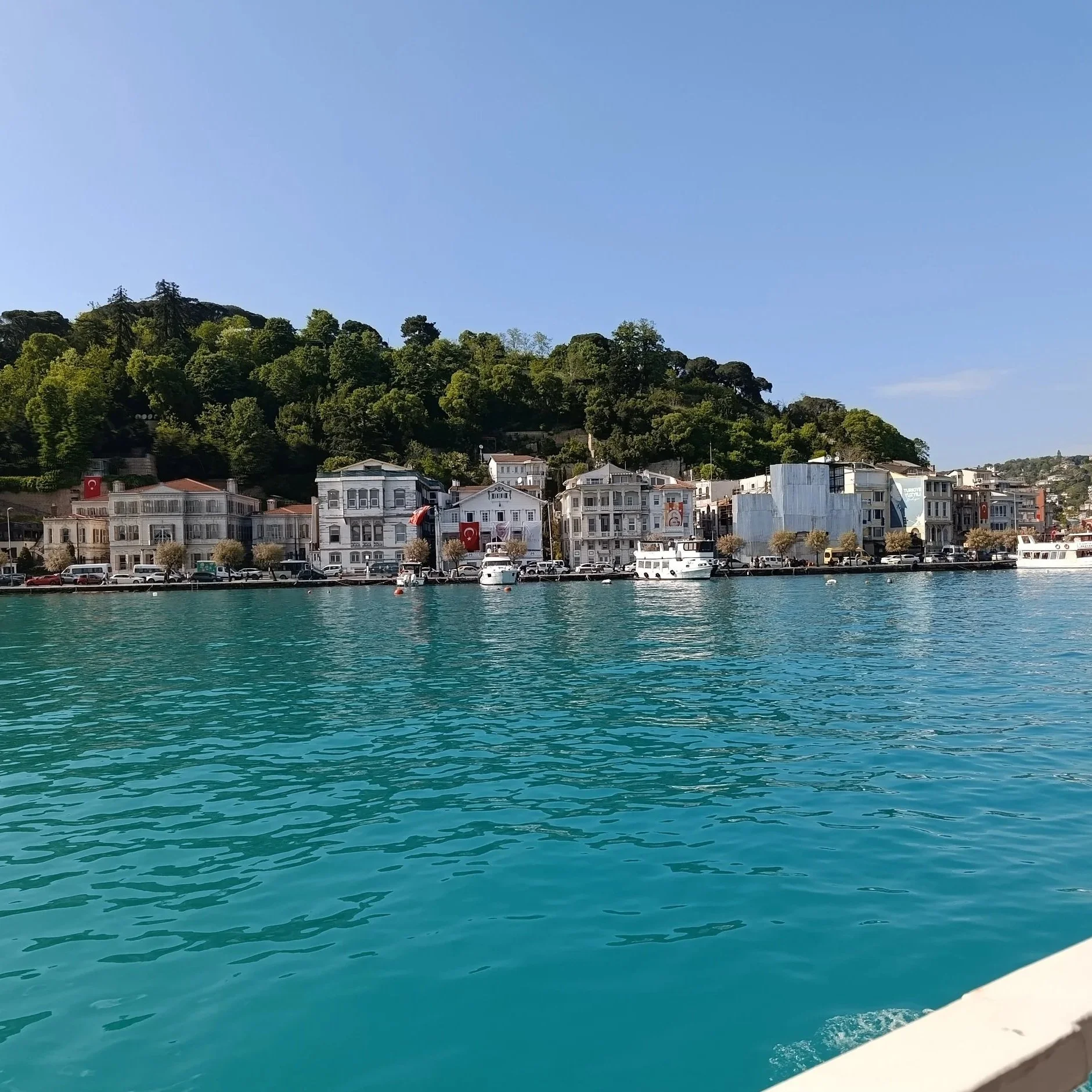 View of a harbor with turquoise water, boats, and waterfront buildings with Turkish flags, set against a lush green hill.
