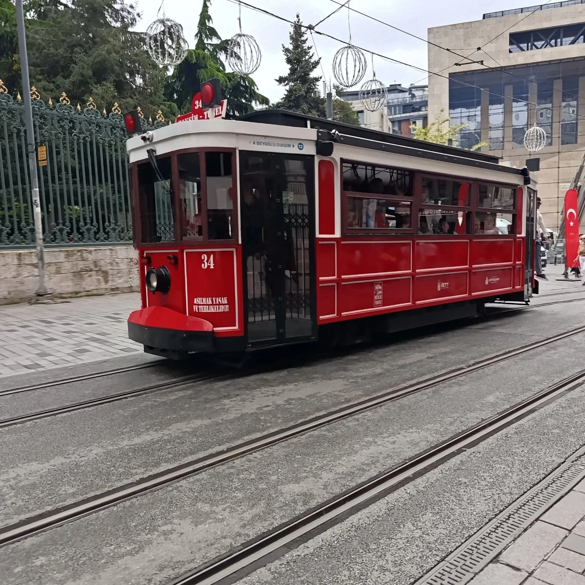 A red tram with the number 34 on it, traveling on tracks in an urban area. The tram has a sign indicating the route from Beyoğlu to Taksim. There are modern buildings and decorative hanging light fixtures above, with trees and a metal fence nearby.