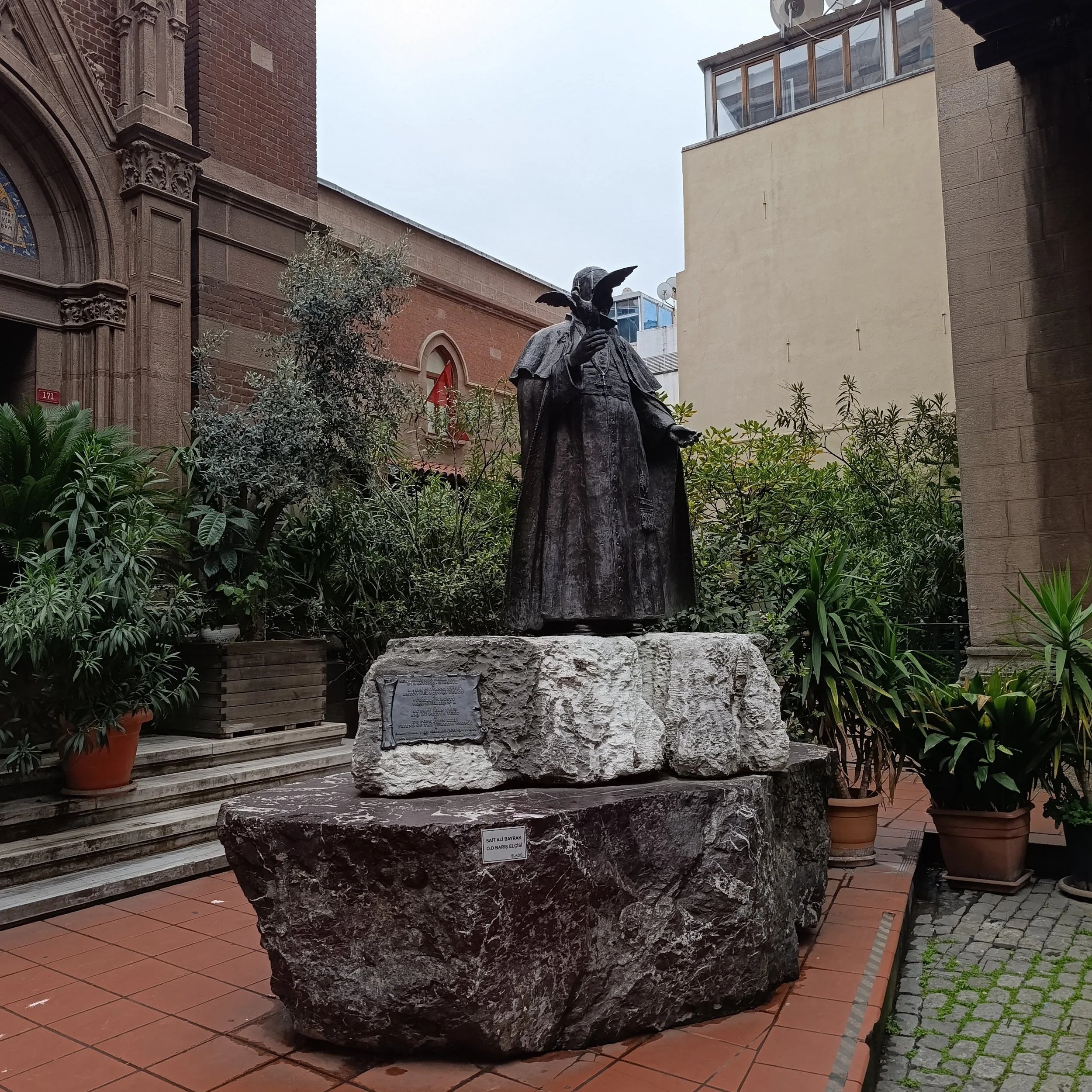 A church building with ornate stone architecture, surrounded by potted plants and trees, with a statue of a girl with wings holding a dove, placed on a large rough rock base in an outdoor courtyard setting.