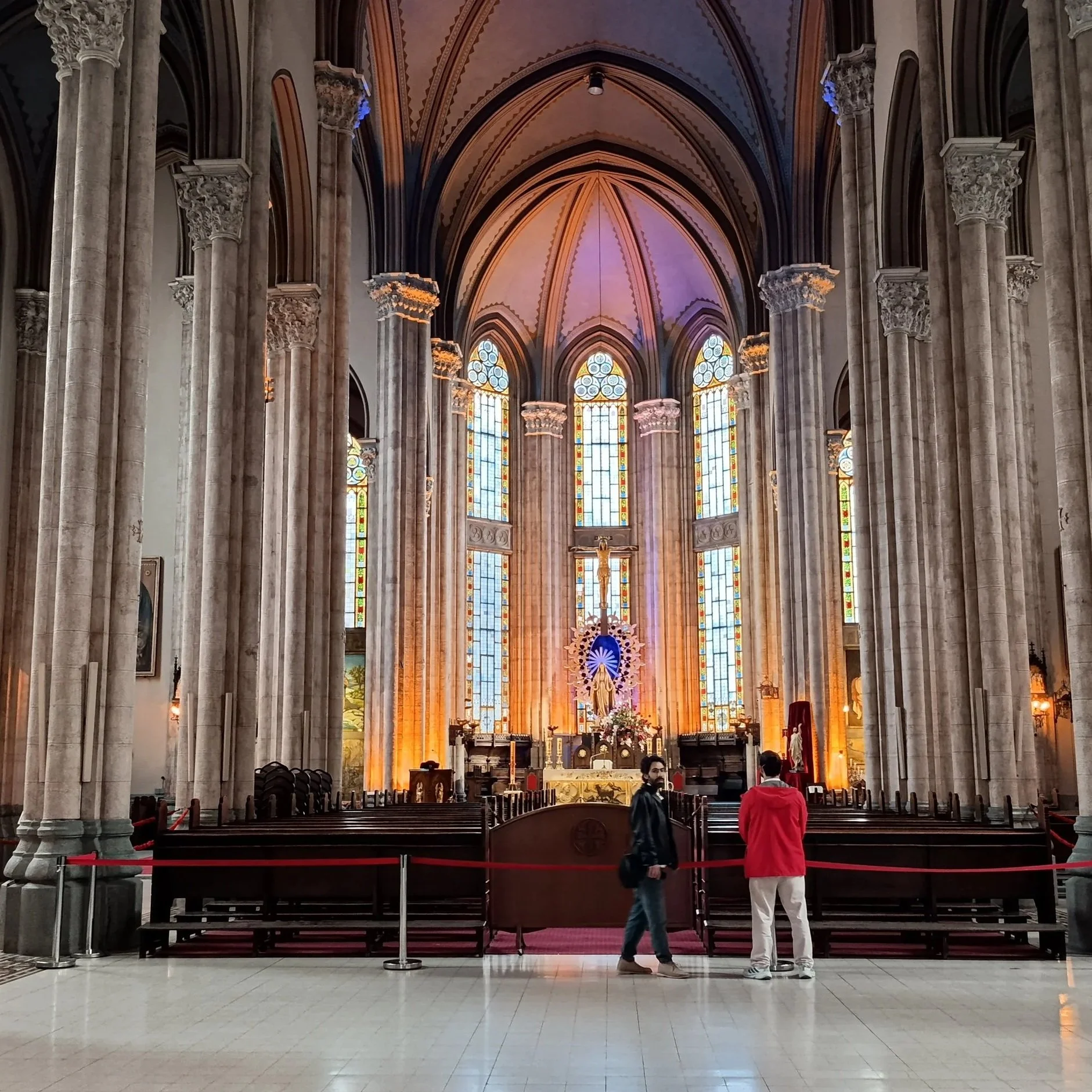 Interior of a Gothic-style church with tall stone columns, stained glass windows, and an altar at the front. Two men are standing near the entrance, one dressed in black and the other in red.