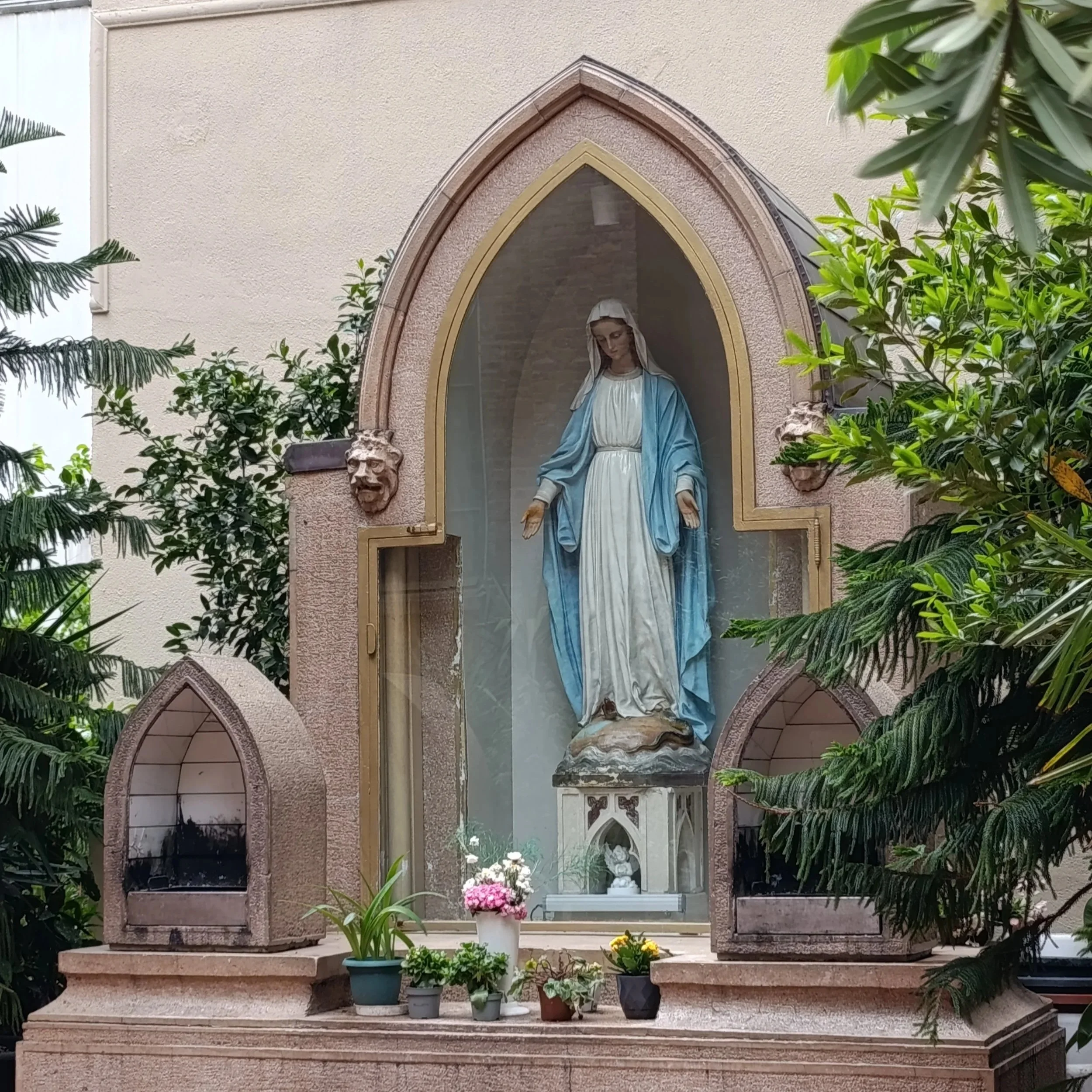 Statue of the Virgin Mary inside a church or religious building, surrounded by green foliage and potted flowers, with stained glass windows behind.