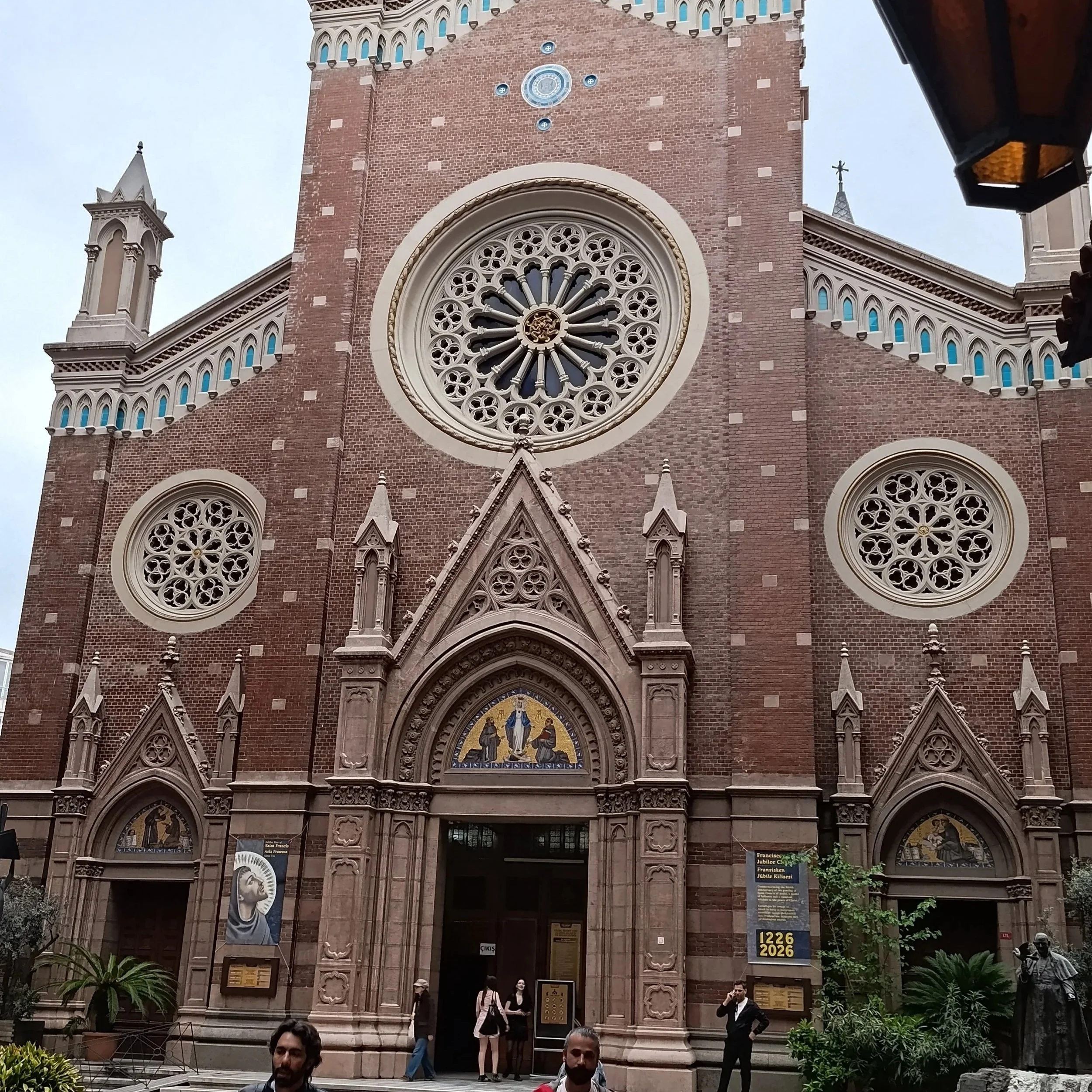 Facade of a large historic church with rose window and arched entrance, people walking and gathered in front, plants and statues near the entrance.
