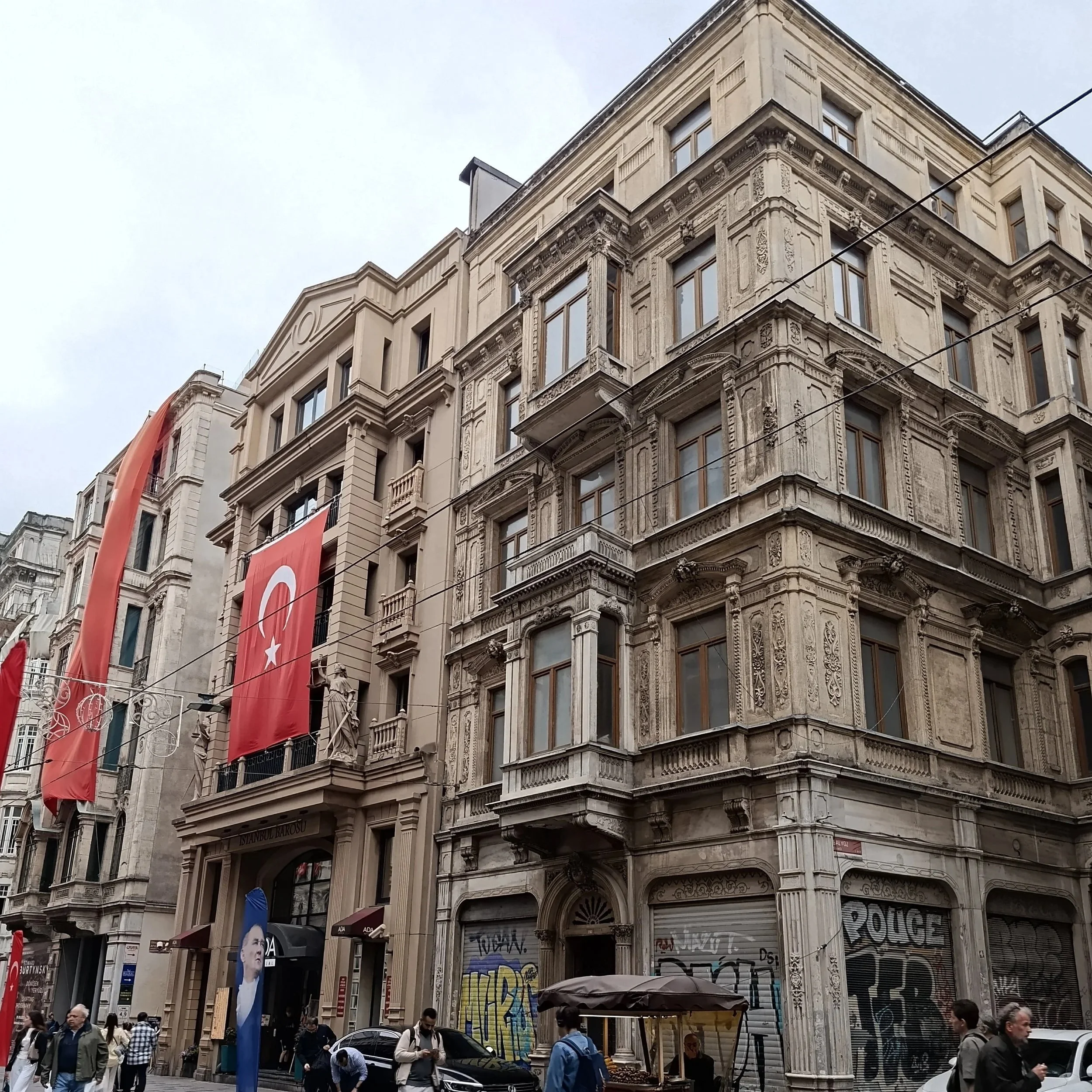 Street view of a historic building with ornate architecture, Turkey flags hanging from the facade, and a crowd of people walking below.