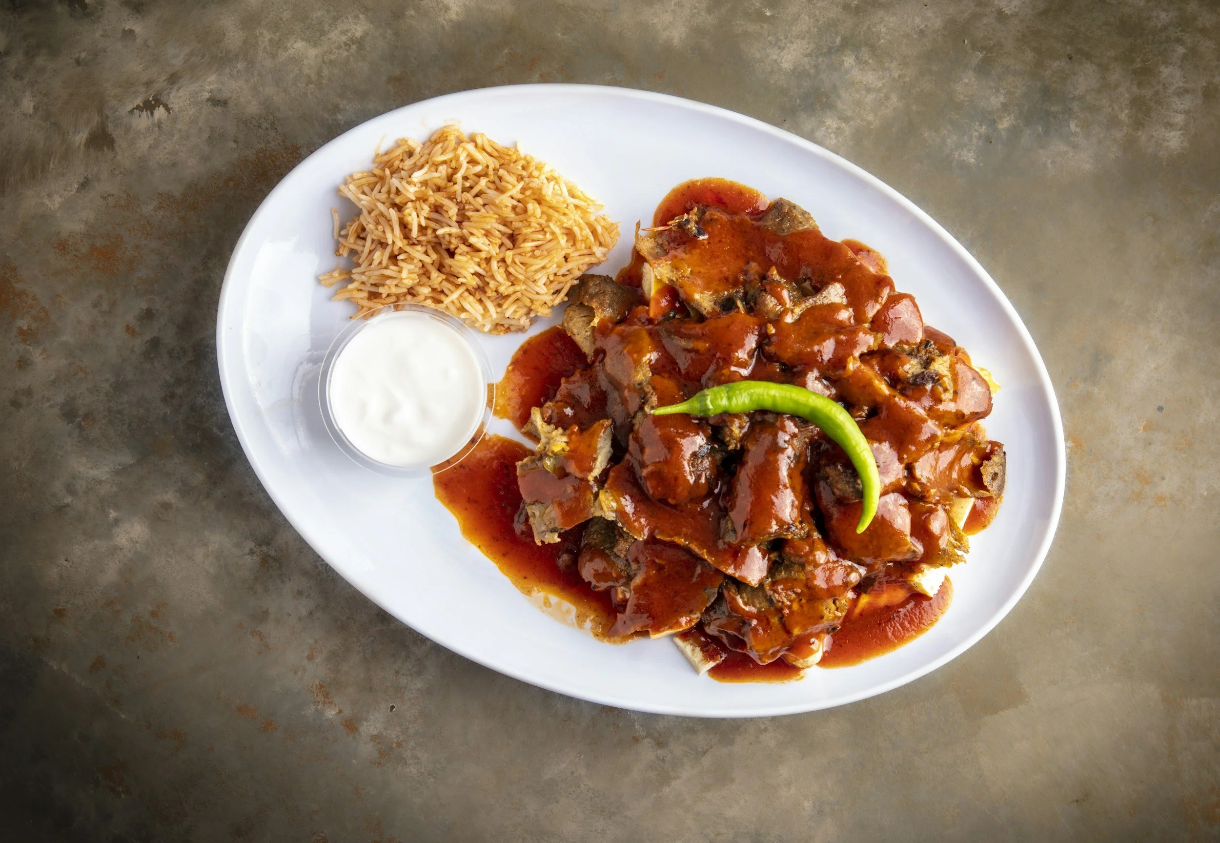 Plate of rice with tomato sauce, meat stew with vegetables, a small cup of white sauce, and a green chili pepper on a gray surface.