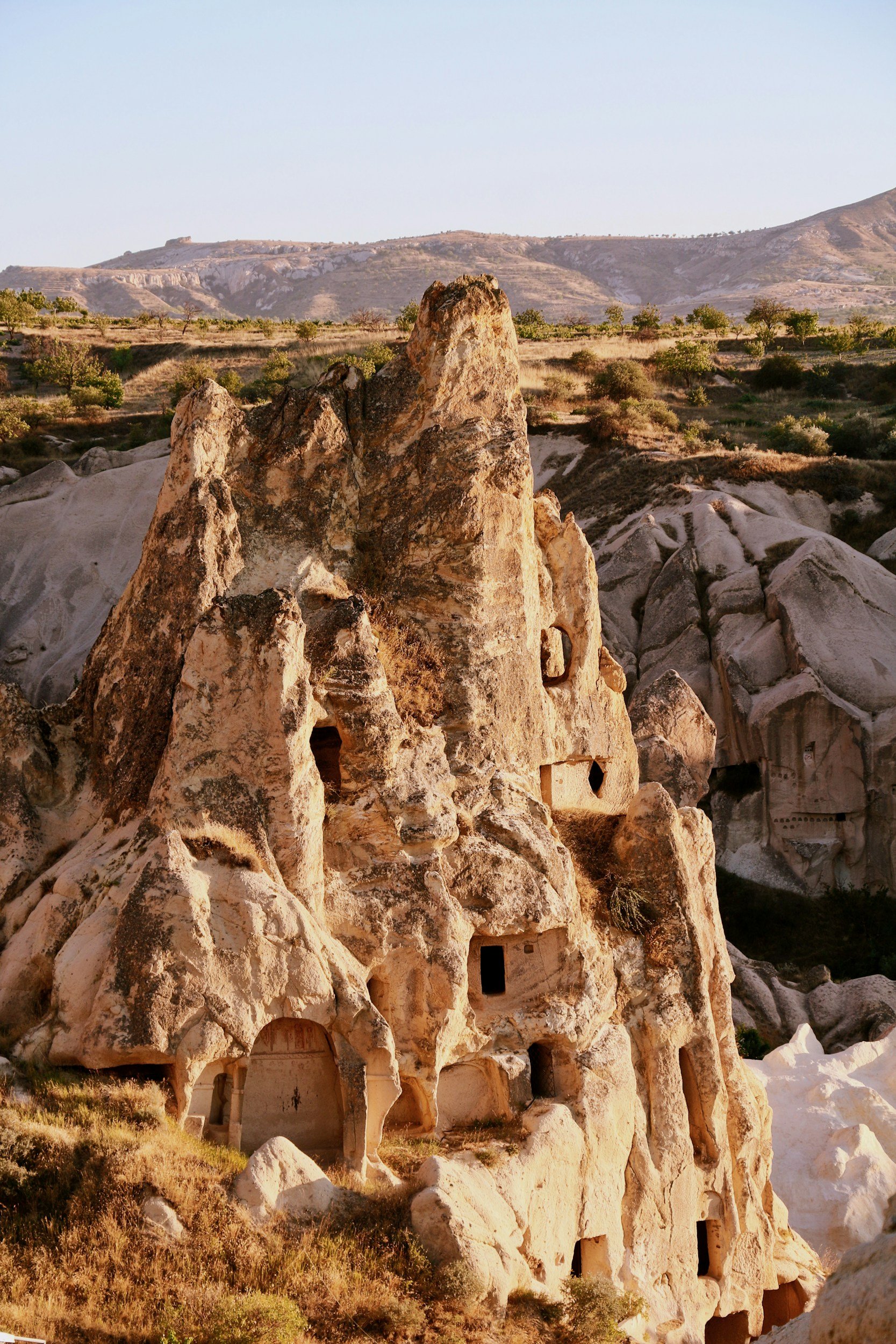 Rock formations with carved dwellings in the cliffs of Cappadocia, Turkey, with distant mountains and clear sky in the background.