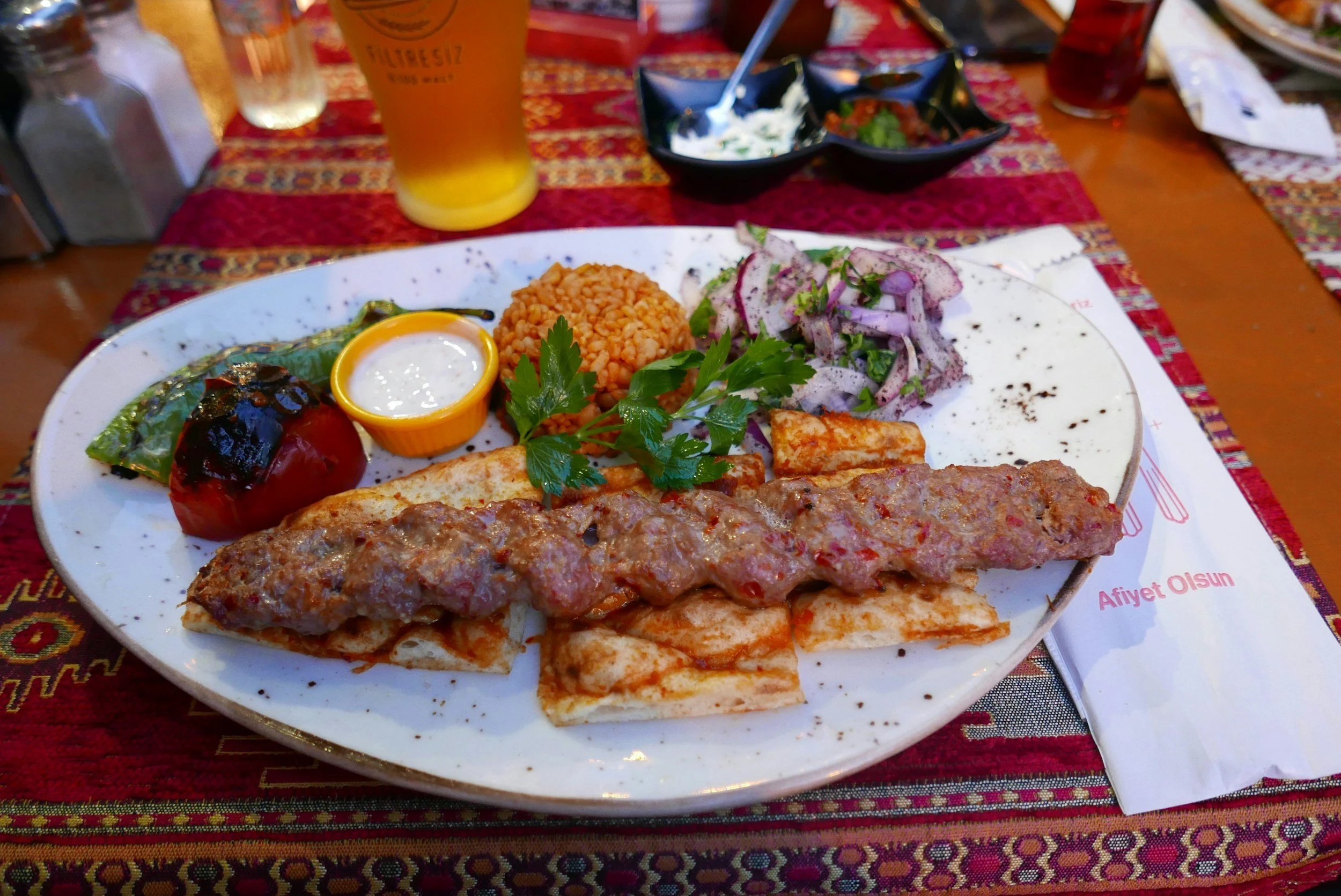 A plate of Turkish food including grilled kebabs, rice, grilled vegetable, salad, and sauces, set on a patterned tablecloth with drinks and bowls in the background.