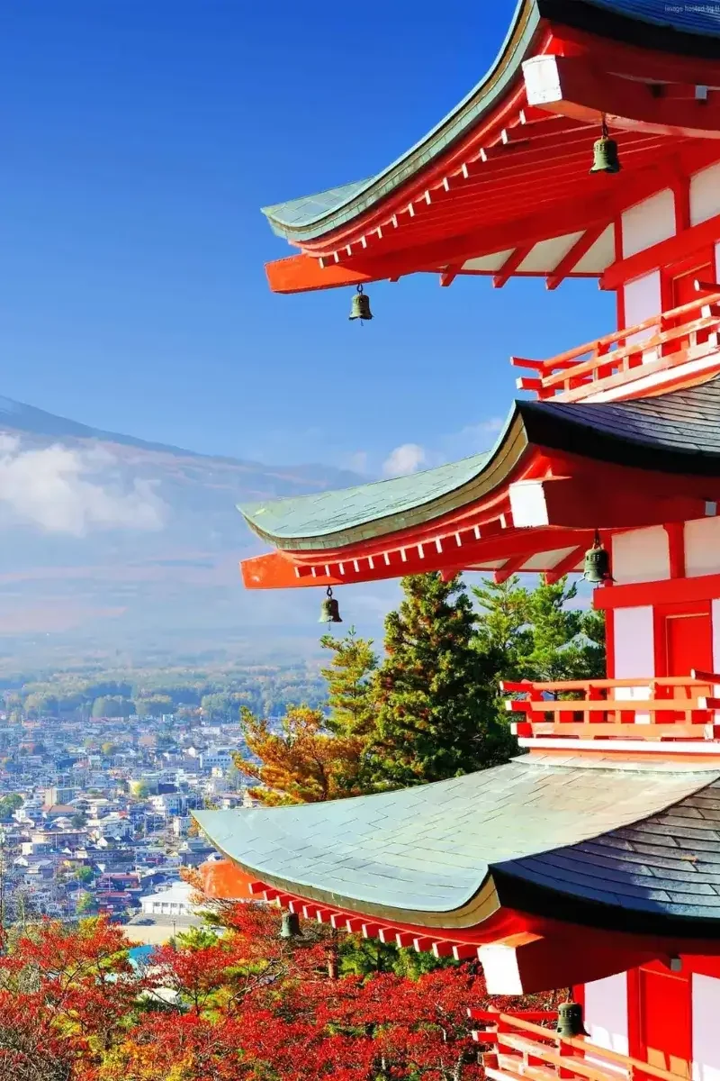 View of a traditional Japanese pagoda with red and white colors, multiple eaves, and small bells hanging from each eave, set against a scenic backdrop of a cityscape and distant mountains during autumn.