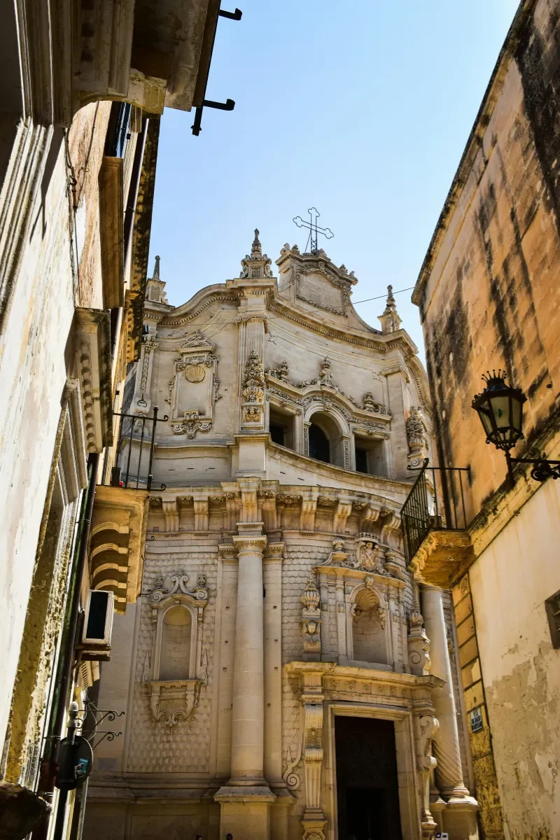View of an ornate historic church with elaborate carvings and a cross on top, seen from a narrow street with old buildings on both sides under a clear blue sky.
