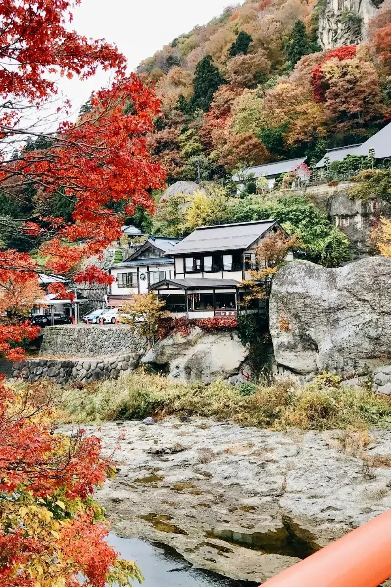 A scenic view of a mountain landscape with trees in fall colors, a river flowing over rocks in the foreground, and traditional Japanese-style houses built on a hillside.