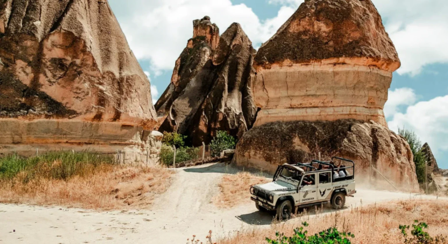 A white off-road vehicle driving on a dirt path among large rock formations and sparse vegetation in a desert landscape.