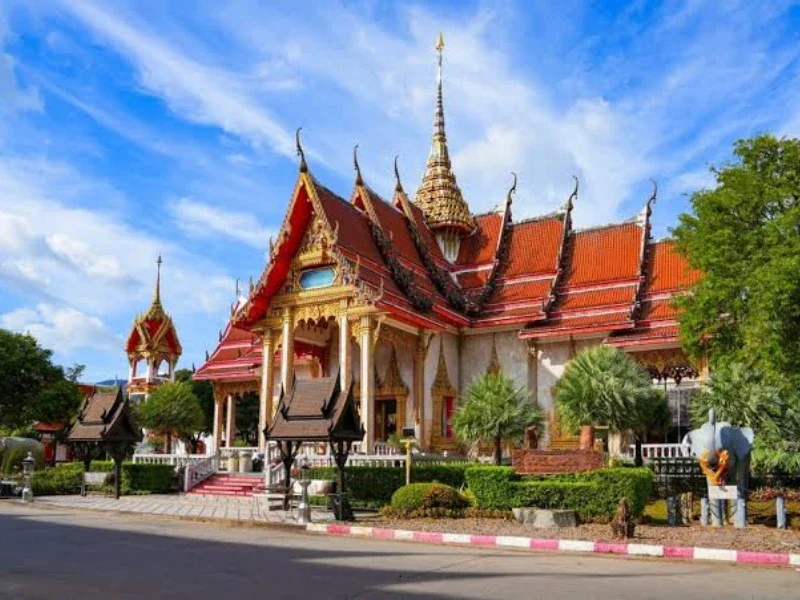 Traditional Thai temple with ornate roof details, surrounded by greenery and a clear blue sky.