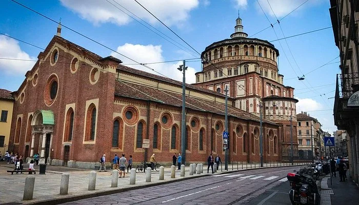 A historic church with red brick walls and rounded windows, situated on a city street with pedestrians and motorcycles, under a partly cloudy sky.