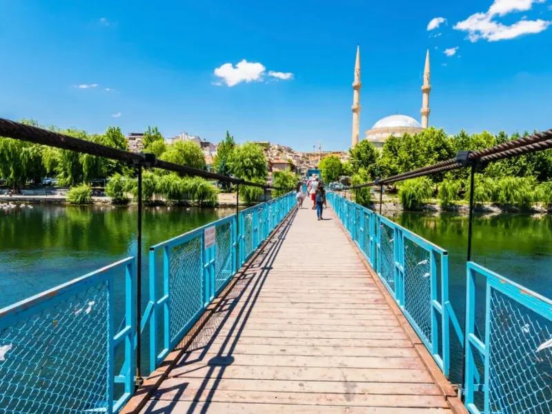 A wooden footbridge with blue railings over a river, with a mosque featuring two minarets in the background, green trees along the riverbanks, and a bright blue sky with scattered clouds.