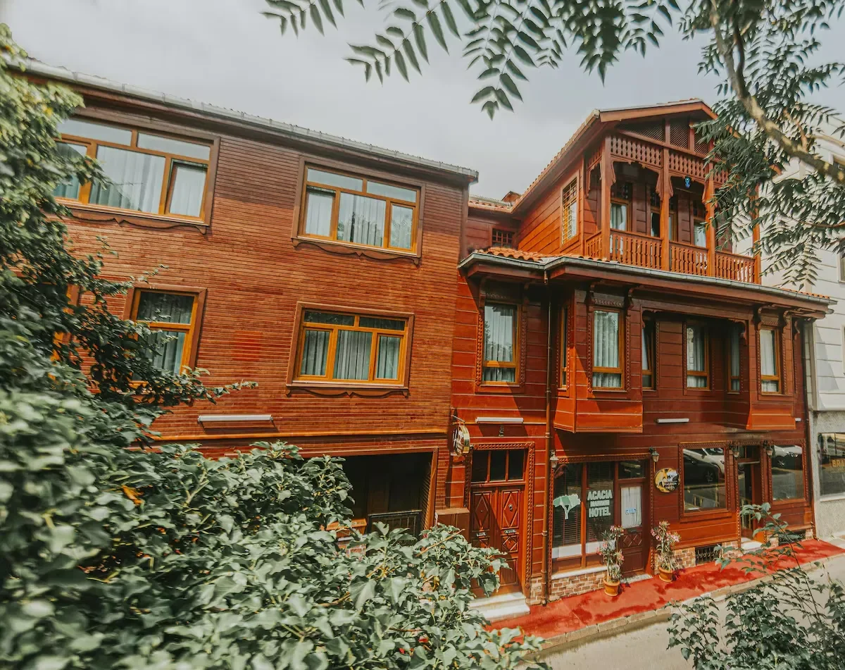 A multi-story wooden building, likely a hotel, with large windows and a small balcony on the upper floor, surrounded by leafy trees and foliage.