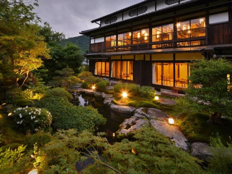 Traditional Japanese-style house with large windows and warm interior lighting, surrounded by a lush garden with rocks, bushes, and trees, illuminated by outdoor lights at dusk.