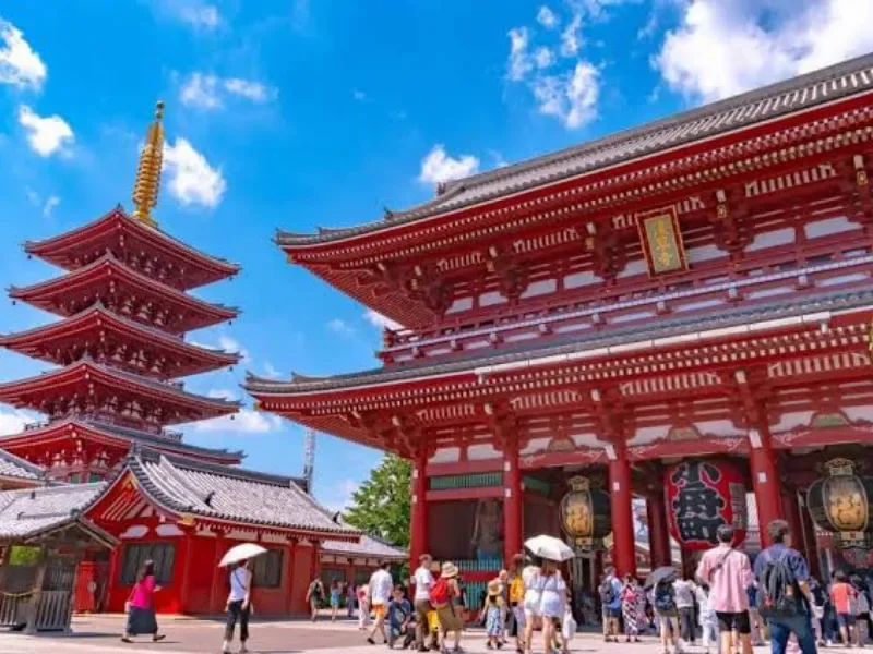 Traditional Japanese temple with a five-story pagoda, red wooden structures, and people tourists visiting outdoors on a sunny day.