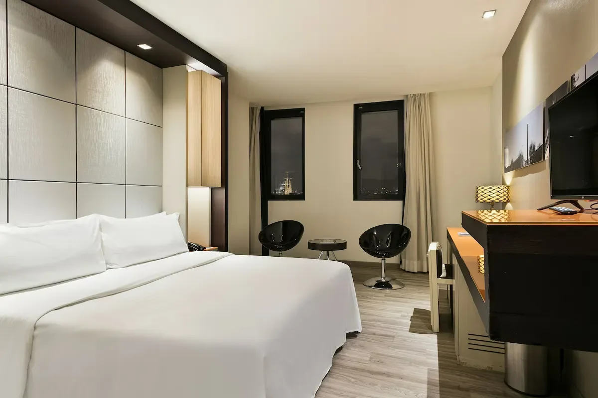 A modern hotel room with a large white bed, two windows, two black chairs with a small table between them, a flat-screen TV, a wooden desk with a lamp, and beige curtains.