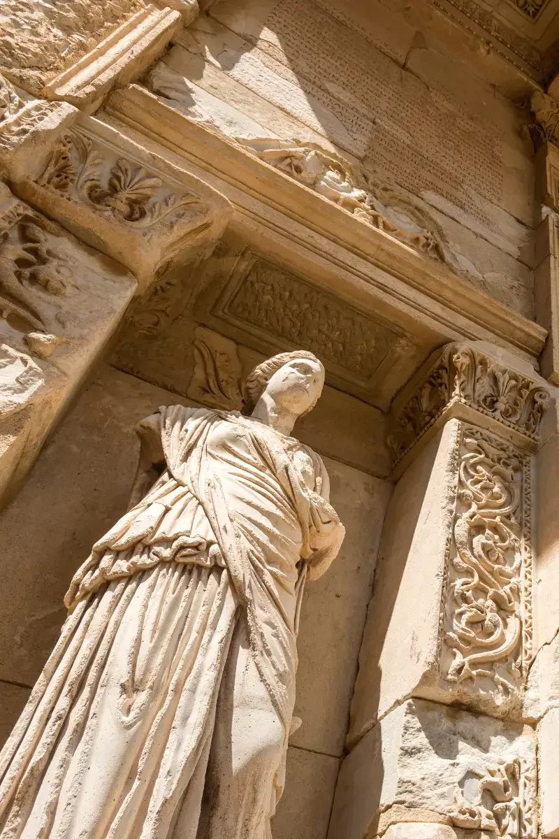 Ancient marble statue of a woman, partly damaged, standing in front of an elaborately decorated stone wall with carved floral patterns.