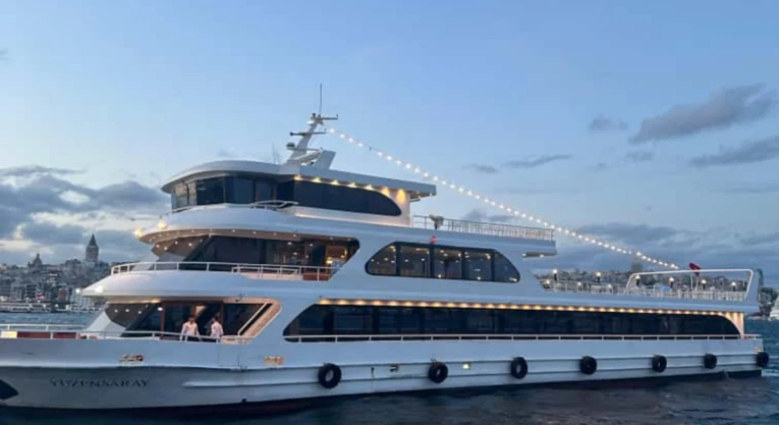 Luxury white yacht docked on the water at dusk, with a city skyline in the background.