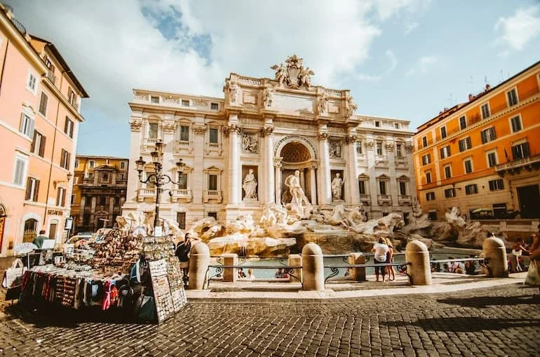 People gather around the Trevi Fountain in Rome, Italy, with vendor stalls on the side selling souvenirs and jewelry, surrounded by historic yellow and orange buildings on a sunny day.