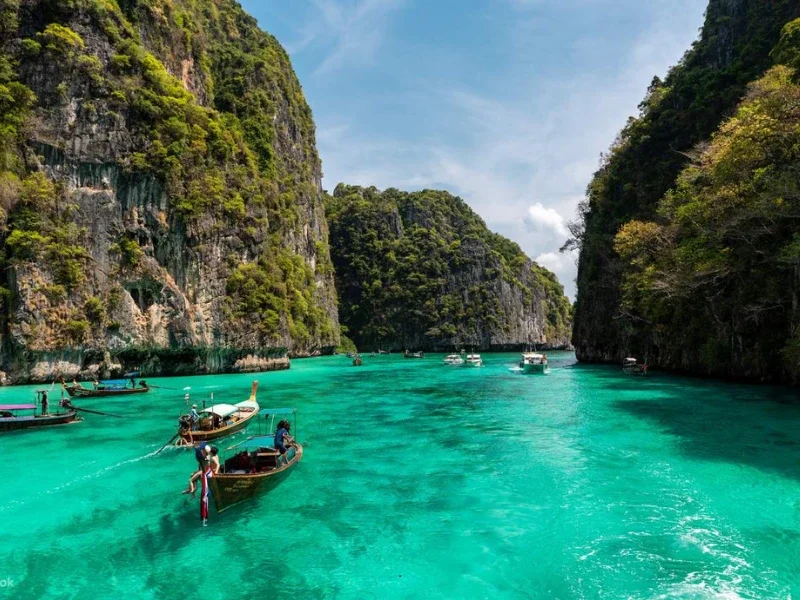 Turquoise waters between steep, lush green cliffs, with several boats floating in the water under a partly cloudy sky.