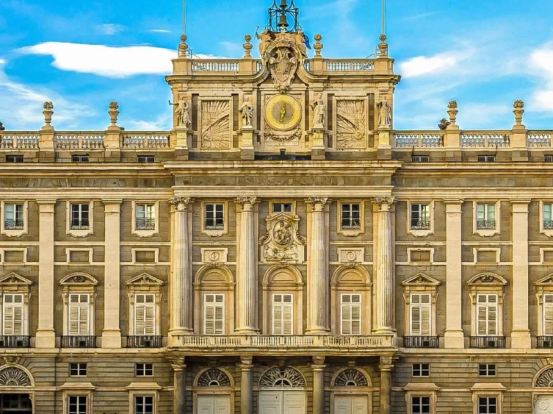 The facade of a historic, ornate building with classical architecture, featuring large columns, decorative sculptures, and a clock at the top center against a blue sky.