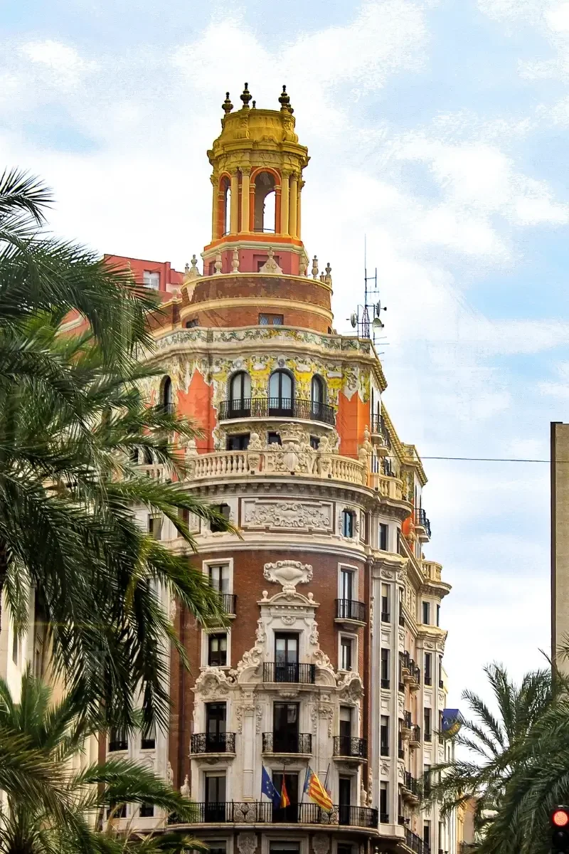 A colorful historic building with ornate architectural details, balconies, and a tower, framed by palm trees and set against a cloudy sky in an urban area.
