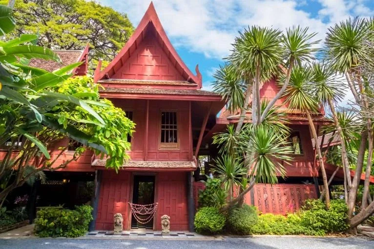 Traditional Thai-style wooden house with a steep gabled roof, surrounded by lush tropical plants and trees.