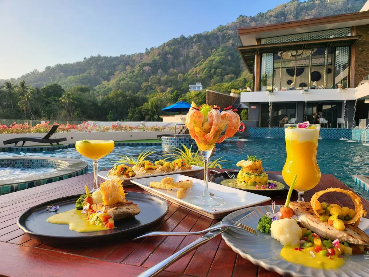 Assorted gourmet dishes and colorful cocktails on a table by a swimming pool, with a modern building and lush green hills in the background.