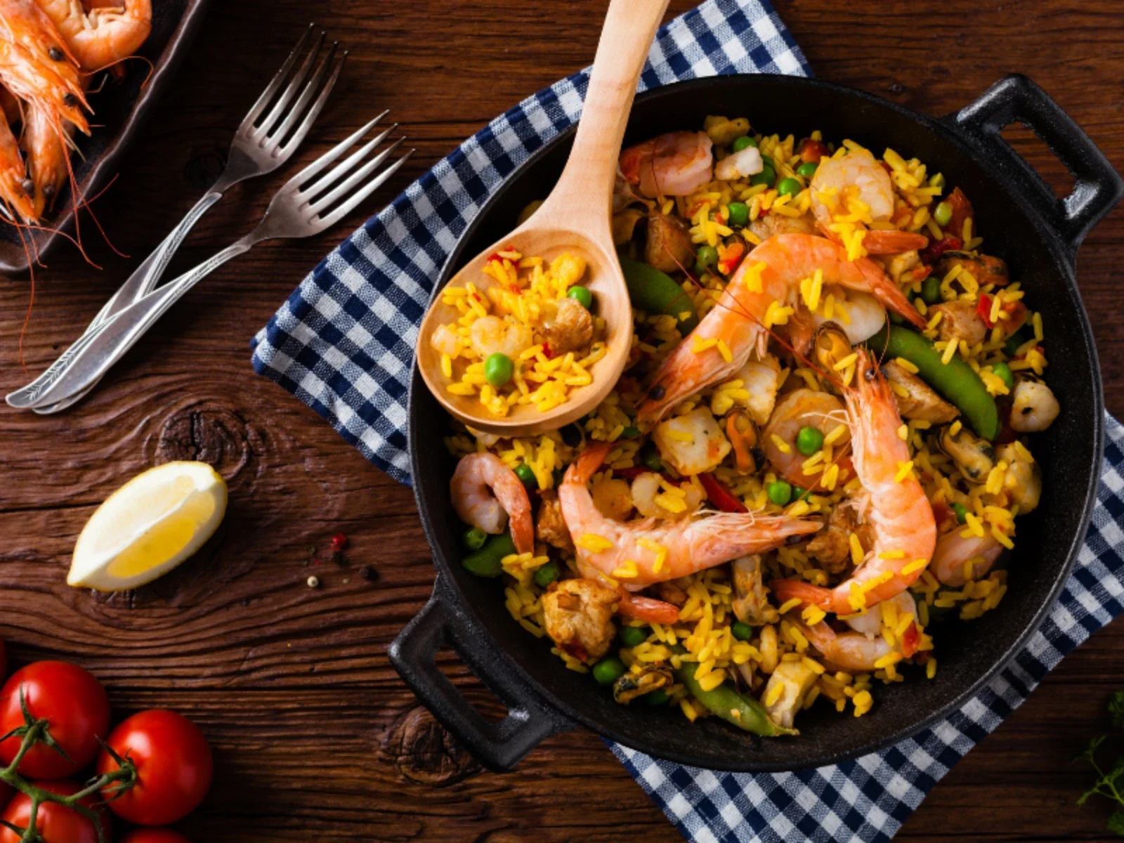 Seafood paella in a black skillet with shrimp, rice, vegetables, and lemon wedge on a wooden table with forks, cherry tomatoes, and a blue checkered cloth.