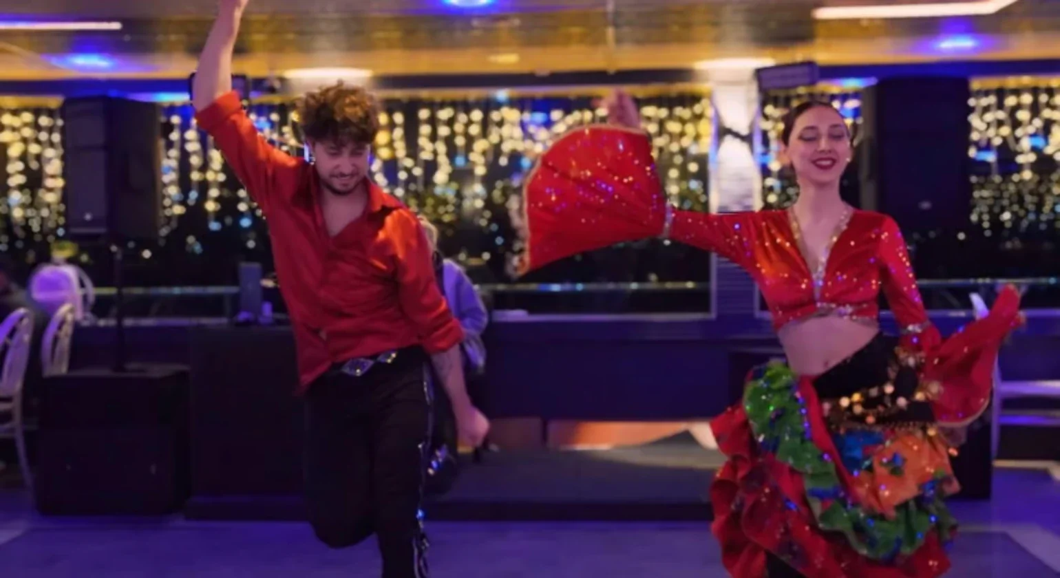A man and a woman dancing indoors at a lively event, with colorful lighting and string lights in the background. The woman wears a vibrant red and orange patterned dress with green accents and holds a matching red fan, while the man wears a red shirt and black pants.