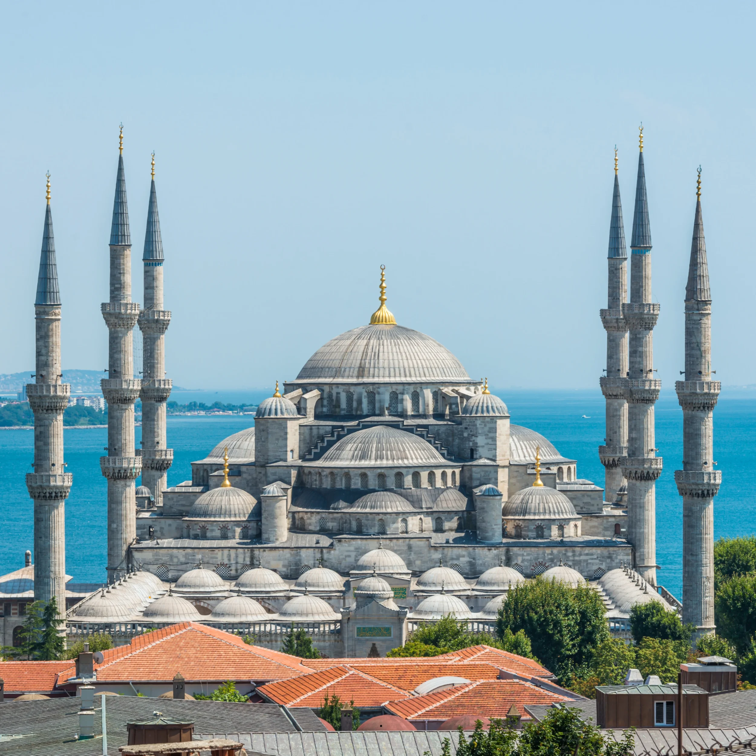 View of a mosque with multiple minarets, domes, and red-tiled roofs, overlooking a water body with a clear sky.