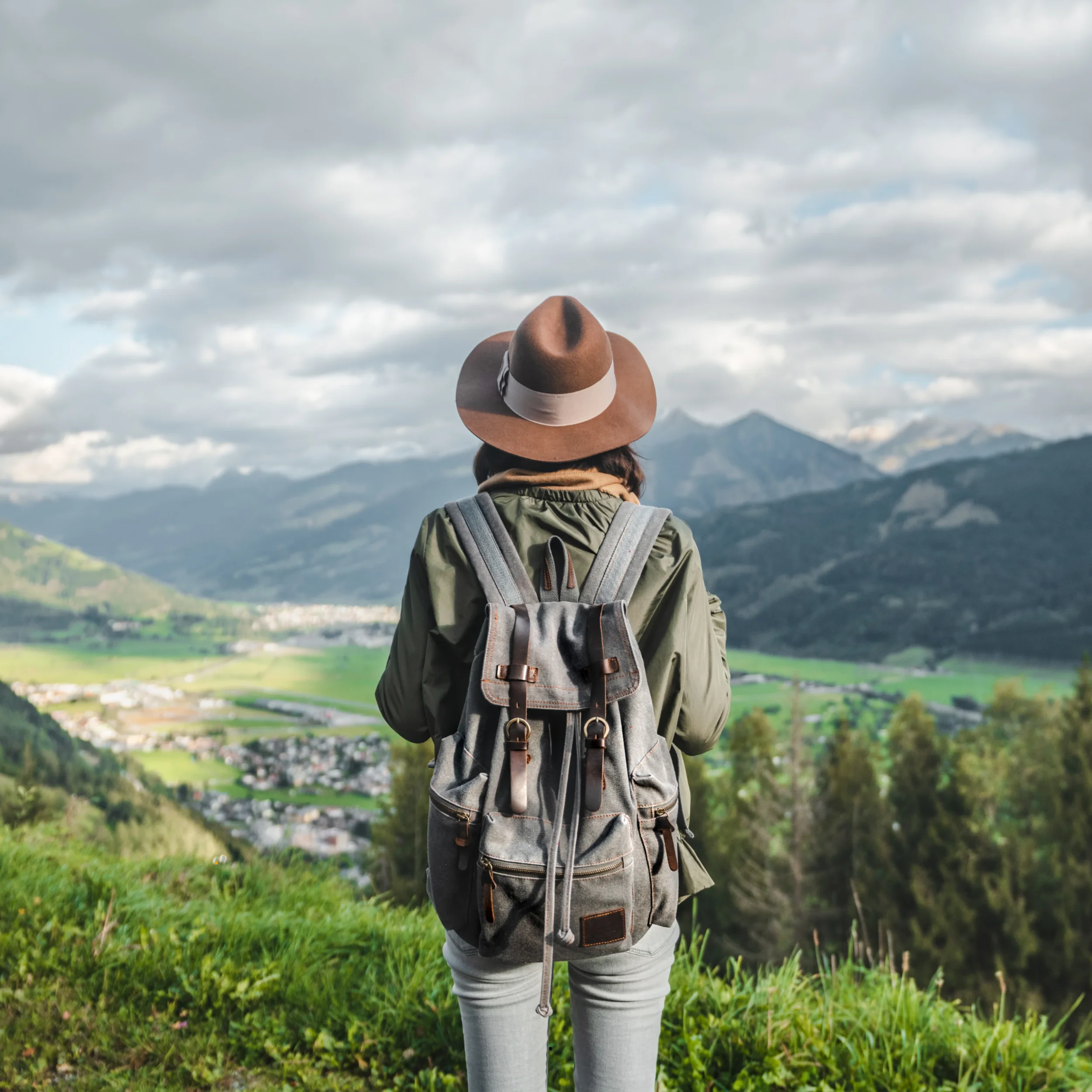 Person wearing a wide-brimmed hat and gray jacket standing outdoors in a green field, with mountains and cloudy sky in the background.